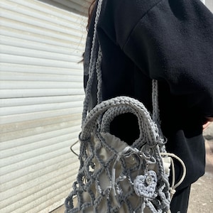 May include: A grey crocheted bag with a white mesh lining and a small grey heart charm. The bag has a long strap and a top handle.
