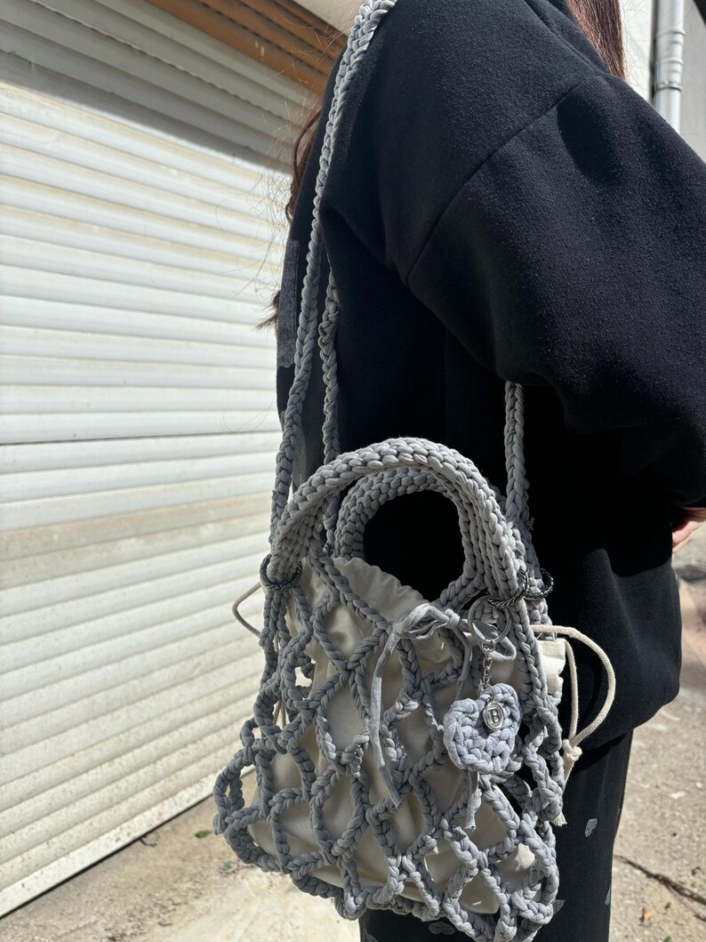 May include: A grey crocheted bag with a white mesh lining and a small grey heart charm. The bag has a long strap and a top handle.