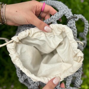 May include: A grey crocheted bag with a white cotton lining. The bag has a drawstring closure and is open to reveal the lining.
