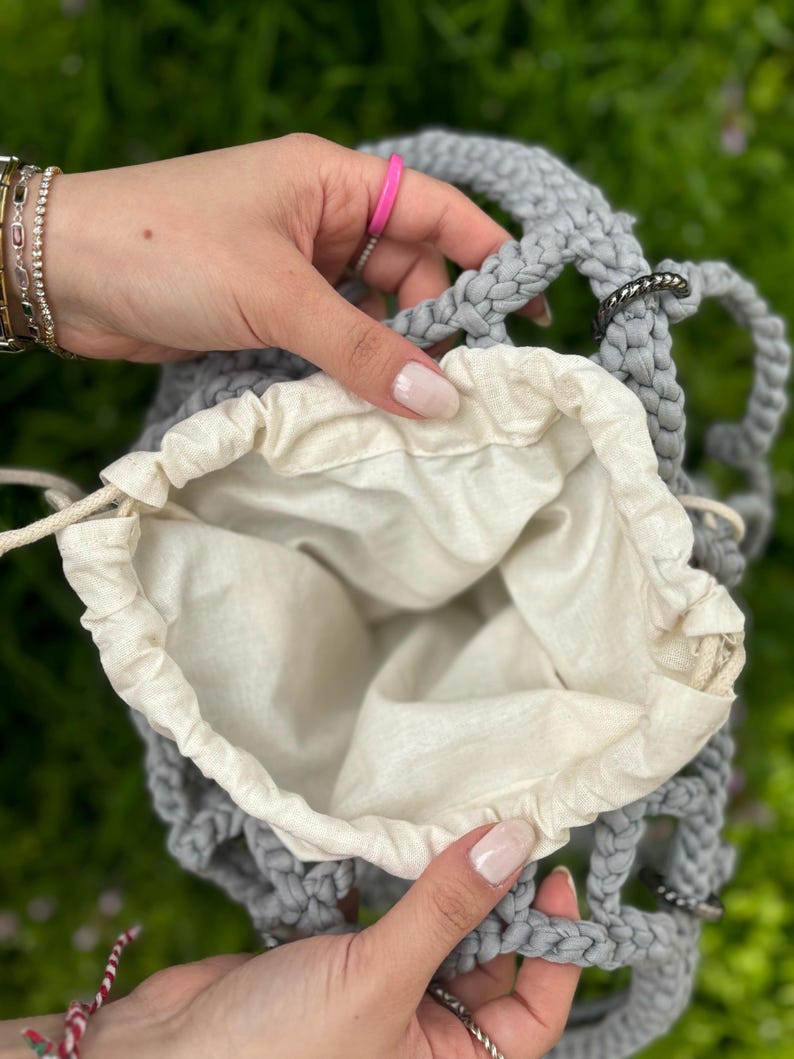 May include: A grey crocheted bag with a white cotton lining. The bag has a drawstring closure and is open to reveal the lining.