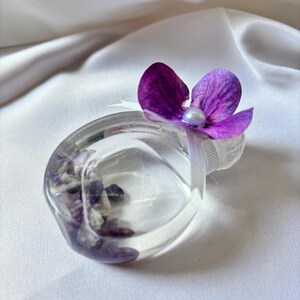 May include: A clear, ring-shaped resin object filled with purple and white stones, topped with a vibrant purple flower and a small white pearl. The object is set against a white, silky background.