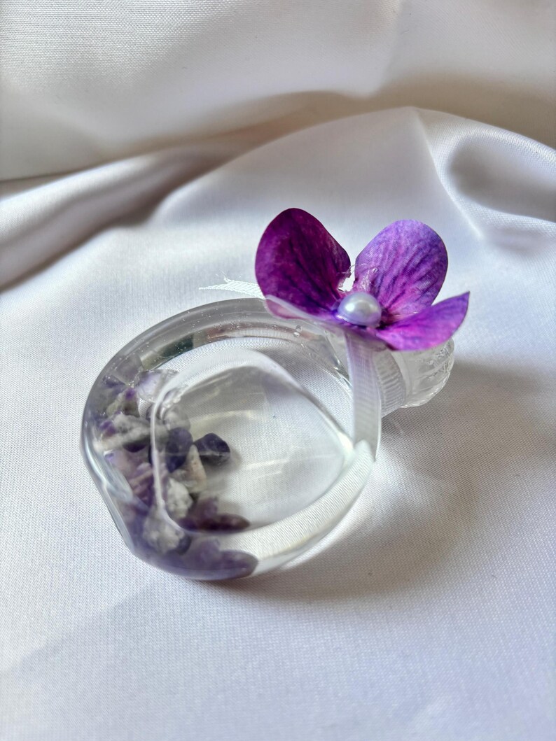 May include: A clear, ring-shaped resin object filled with purple and white stones, topped with a vibrant purple flower and a small white pearl. The object is set against a white, silky background.
