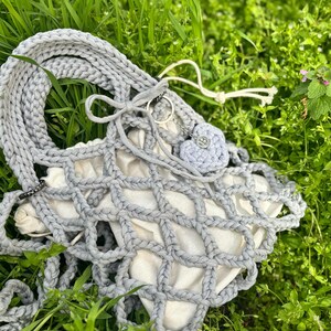 May include: A grey crocheted tote bag with a white mesh lining. The bag has a long strap and a small grey crocheted heart charm.