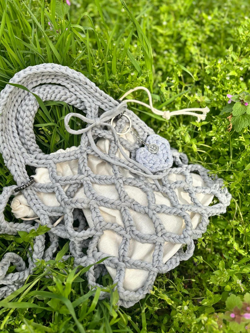 May include: A grey crocheted tote bag with a white mesh lining. The bag has a long strap and a small grey crocheted heart charm.