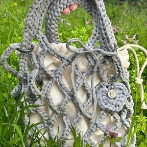 May include: A grey crocheted tote bag with a white mesh lining and a grey crocheted heart charm with the letter "B" on it.