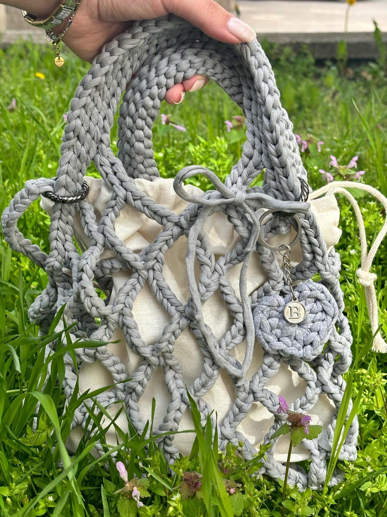May include: A grey crocheted tote bag with a white mesh lining and a grey crocheted heart charm with the letter "B" on it.