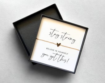 Könnte beinhalten: Ein goldfarbenes Armband mit Herzanhänger in einer schwarzen Geschenkbox. Die Box enthält eine weiße Karte mit den Sätzen "stay strong", "believe in yourself" und "you got this!" in schwarzer Schrift.