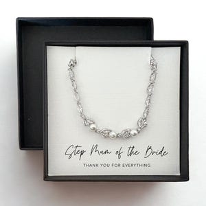 Puede incluir: Un brazalete plateado con detalles de perlas descansa en una caja de regalo negra. El brazalete se exhibe en una tarjeta blanca con el texto "Step Mum of the Bride" y "Thank you for everything". La caja está abierta.
