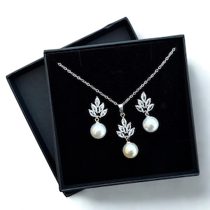 May include: A silver necklace and earring set with a leaf design and white pearls, presented in a black box. The necklace has a central pendant and the earrings match. The set is displayed on a black background, creating a contrast with the jewellery.