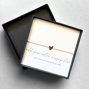 May include: A gold-colored heart pendant necklace displayed in an open black gift box. The necklace is on a white card with the text "add your custom message here". The box is on a light gray surface.
