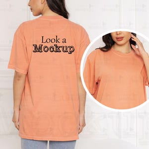 Puede incluir: Camiseta holgada de color melocotón con el texto "Look a Mockup" impreso en negro en la espalda. La camiseta tiene mangas cortas y cuello redondo. La modelo lleva vaqueros azules.
