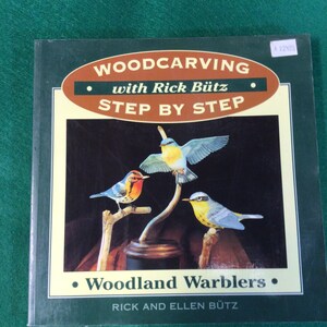 Peut inclure: Un livre intitulé "Woodcarving with Rick Bütz Step by Step" présente une illustration de trois parulines des bois colorées perchées sur des branches. La couverture est vert foncé avec du texte et un design ovale marron.