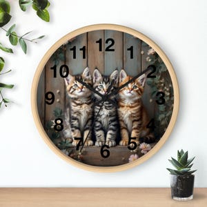 May include: A round wall clock with a light wood frame. The clock face features three kittens with brown and black tabby markings, set against a wooden plank background. Black numerals mark the hours, and the clock is surrounded by floral accents.