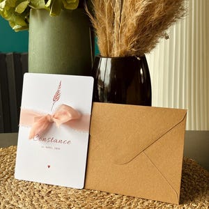 May include: A white invitation card with the name "Constance" and the date "24 AVRIL 2026" in burgundy script, tied with a pink ribbon. A brown envelope rests beside it. The card features a small floral design and a heart symbol.