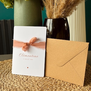 May include: A white invitation card with the name "Clémentine" in brown script, tied with a peach ribbon. A brown envelope sits beside the card on a woven mat. Decorative vases with flowers are in the background.