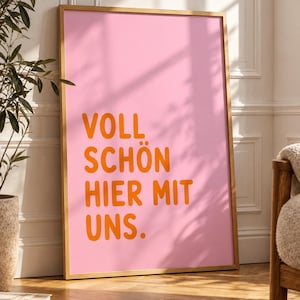 May include: A rectangular wall art print in a light wooden frame featuring a solid pink background with the orange text VOLL SCHÖN HIER MIT UNS. The print is displayed in a vertical orientation, showcasing bold, sans-serif typography.