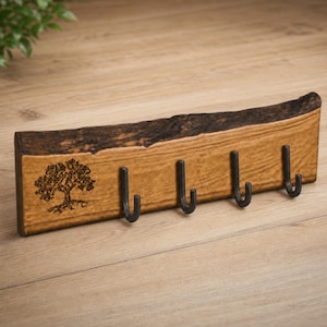 May include: A rectangular wooden wall hook rack featuring a natural live edge top and a dark brown finish. The piece includes four black metal J-shaped hooks and a black laser-engraved illustration of a tree on the left side of the front face.