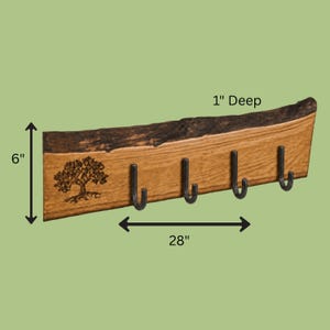 May include: A live edge wooden wall rack with four black metal hooks and a laser-engraved tree design. The board measures 28 inches wide, 6 inches high, and 1 inch deep, featuring a natural bark top edge and a light brown wood grain finish.