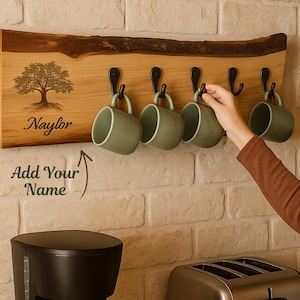 May include: A live edge wooden wall rack featuring five black metal hooks, a laser-engraved tree illustration, and the text "Naylor" and "Add Your Name". Four matte sage green ceramic mugs hang from the hooks on the horizontal wooden board.