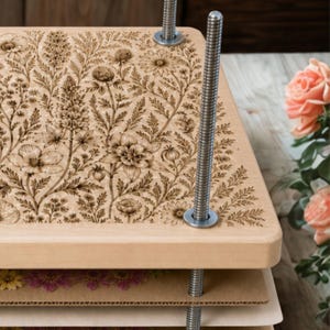 May include: A wooden flower press featuring a top board with an intricate laser-engraved botanical pattern of wildflowers and ferns. The press includes four threaded metal rods with washers and nuts, and several layers of cardboard inserts.