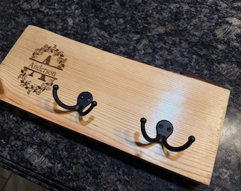May include: A wooden coat rack with two black metal hooks. The light-colored wood has a natural grain and a live edge. The name "Anderson" is engraved above the hooks, surrounded by a floral design.