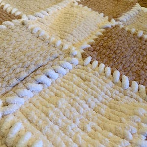 May include: A handmade, plush blanket in a patchwork design. The blanket features squares of cream, tan, and brown, with white stitching. The soft, textured material is visible in this close-up shot of the cozy, warm blanket.