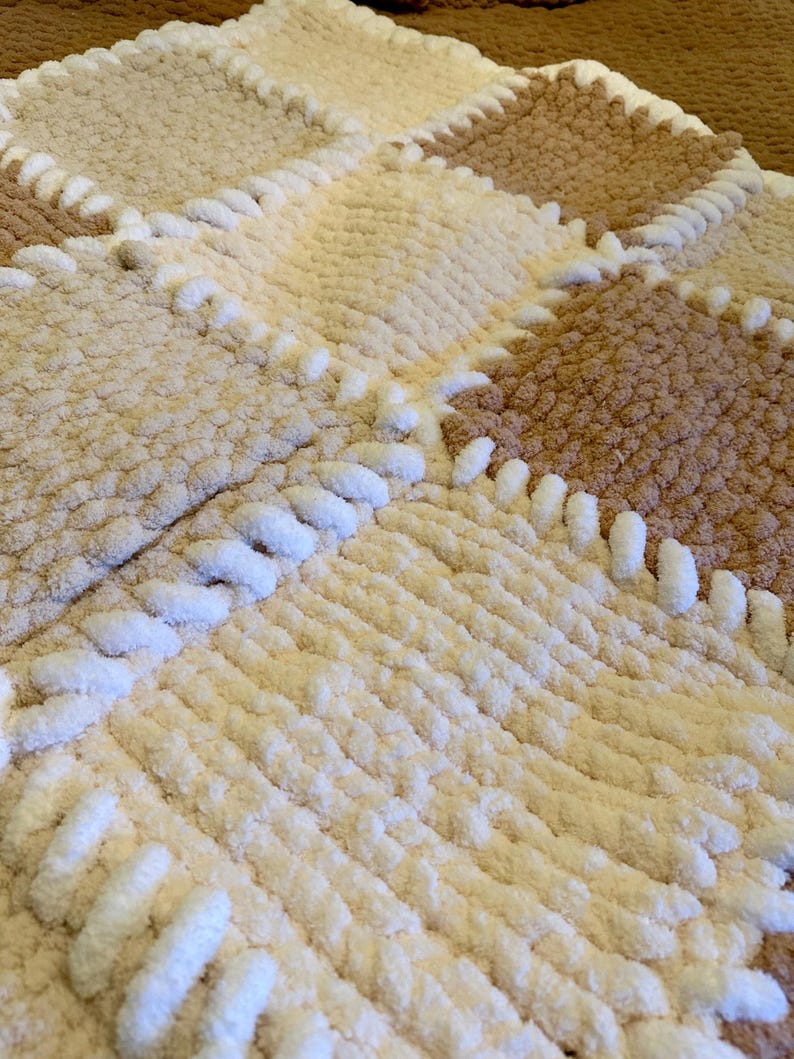 May include: A handmade, plush blanket in a patchwork design. The blanket features squares of cream, tan, and brown, with white stitching. The soft, textured material is visible in this close-up shot of the cozy, warm blanket.