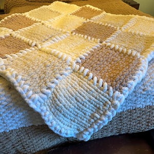 May include: A handmade, square, chunky knit blanket in a patchwork design. The blanket features alternating squares of cream, tan, and light brown. The edges are finished with a thick, white, looped border. The blanket is folded over another blanket.