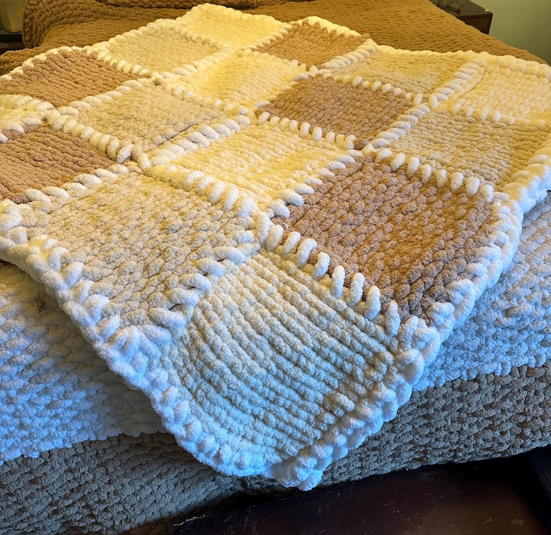 May include: A handmade, square, chunky knit blanket in a patchwork design. The blanket features alternating squares of cream, tan, and light brown. The edges are finished with a thick, white, looped border. The blanket is folded over another blanket.