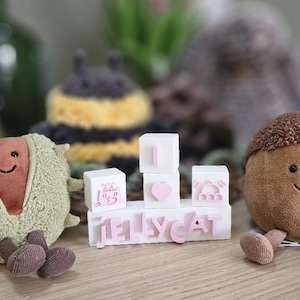 Può includere: Una collezione di peluche, tra cui una castagna, una ghianda e un'ape, con facce sorridenti. I giocattoli sono disposti con blocchi bianchi che compongono "I LOVE Jellycat" in lettere rosa. I giocattoli sono su una superficie di legno.