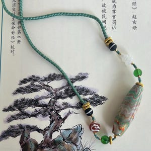 Könnte beinhalten: Eine grüne Schnurkette mit einer großen, ovalen Achatperle mit braunen, weißen und grünen Wirbeln. Die Perle ist von kleineren Perlen in Gold, Schwarz, Weiß und Grün umgeben. Die Kette wird auf einem weißen Hintergrund mit einer Schwarzweiß-Tintenzeichnung einer Kiefer gezeigt.