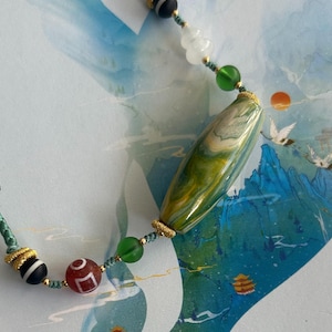 Könnte beinhalten: Eine Halskette mit einem Anhänger aus grünem und weißem Achat mit goldenen Akzenten und grünen, weißen und schwarzen Perlen. Die Halskette ist an einem grünen Band.