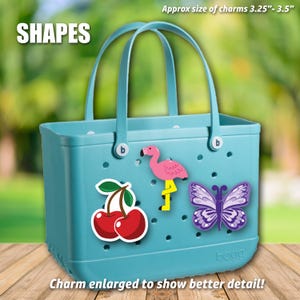 May include: A turquoise tote bag with a flamingo, butterfly, and cherry charms. The bag has two handles and the word "SHAPES" is at the top. The charms are approximately 3.25"-3.5" in size.