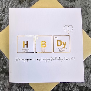 May include: A white square greeting card featuring gold foil periodic table elements for H, B, and Dy, spelling HBD. The card includes a small balloon illustration and the printed text, Wishing you a very Happy Birthday Hannah!