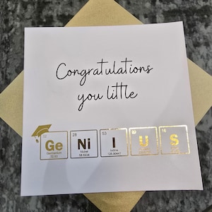 Puede incluir: Tarjeta de graduación blanca con las palabras "Congratulations you little" en escritura negra. La tarjeta presenta elementos dorados de la tabla periódica que deletrean "Genius". La tarjeta está sobre un sobre dorado.