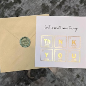 Peut inclure: Une carte carr&eacute;e blanche avec des &eacute;l&eacute;ments du tableau p&eacute;riodique en feuille d'or &eacute;pelant THANK YOU et le texte Just a small card to say, accompagn&eacute;e d'une enveloppe beige clair avec un sceau de cire vert portant le texte Open me.