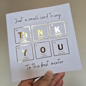 Peut inclure: Une carte de v&oelig;ux carr&eacute;e blanche avec le texte Just a small card to say en haut et To the best mentor en bas. Le centre affiche le mot THANK YOU en utilisant des &eacute;l&eacute;ments du tableau p&eacute;riodique en feuille d'or pour chaque lettre.