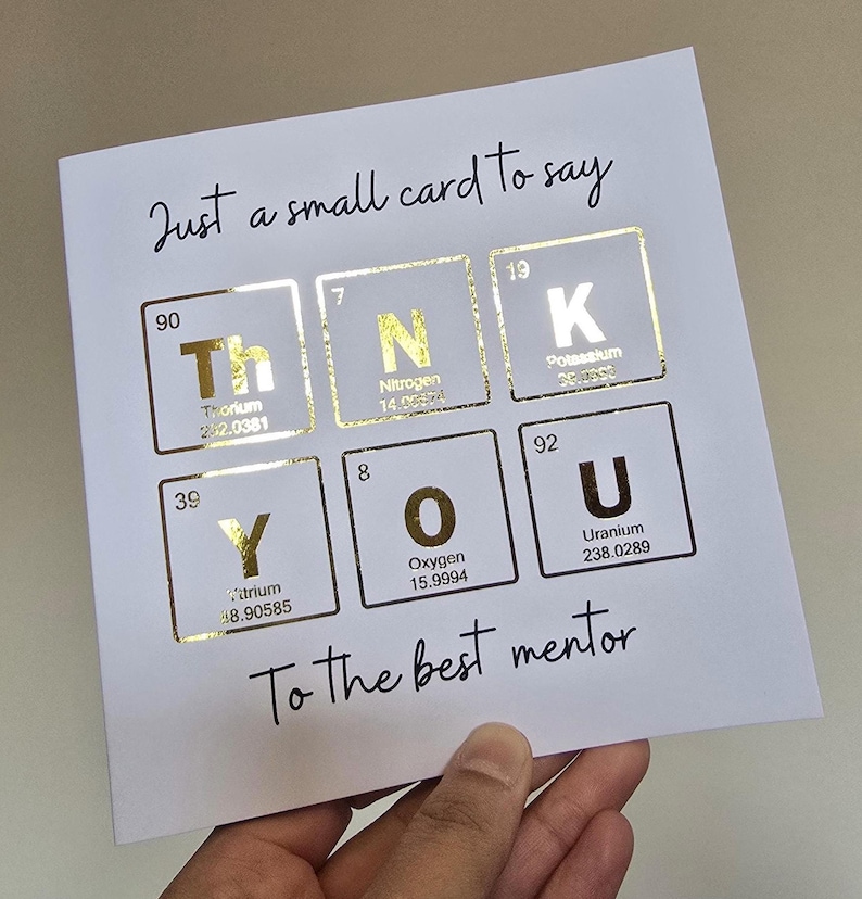 Peut inclure: Une carte de v&oelig;ux carr&eacute;e blanche avec le texte Just a small card to say en haut et To the best mentor en bas. Le centre affiche le mot THANK YOU en utilisant des &eacute;l&eacute;ments du tableau p&eacute;riodique en feuille d'or pour chaque lettre.