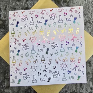 May include: White greeting card with a repeating pattern of science-themed illustrations in black, with colourful holographic accents. The card features images of beakers, atoms, and chemical formulas. The card is on a gold envelope.