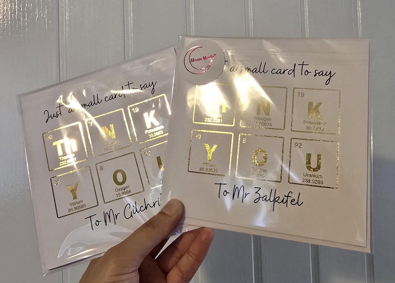 Peut inclure: Deux cartes de v&oelig;ux carr&eacute;es blanches sous pochettes plastiques transparentes avec des &eacute;l&eacute;ments du tableau p&eacute;riodique en feuille d'or formant THANK YOU. Le texte indique Just a small card to say THANK YOU To Mr Gilchrist et To Mr Zalkifel.