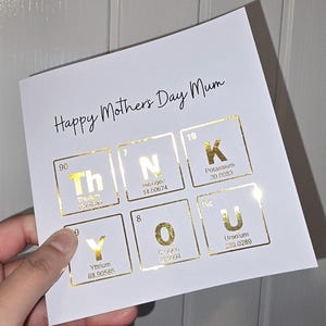 Peut inclure: Une carte de v&oelig;ux carr&eacute;e blanche avec le texte noir Happy Mothers Day Mum en haut. En dessous, six &eacute;l&eacute;ments du tableau p&eacute;riodique en feuille d'or &eacute;pellent THANK YOU, avec les symboles Th, N, K, Y, O, U, leurs num&eacute;ros et noms atomiques.