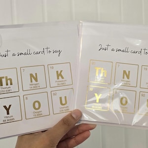 Peut inclure: Deux cartes de v&oelig;ux carr&eacute;es blanches dans des pochettes en plastique transparent. Chaque carte pr&eacute;sente le texte Just a small card to say et un design de tableau p&eacute;riodique en feuille d'or &eacute;pelant THANK YOU avec les &eacute;l&eacute;ments Thorium, Azote, Potassium, Yttrium, Oxyg&egrave;ne et Uranium.