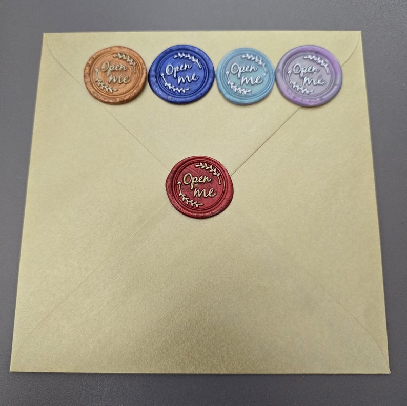 Peut inclure: Une enveloppe carr&eacute;e beige orn&eacute;e de cinq sceaux de cire circulaires. Quatre sceaux sont align&eacute;s en haut et un est centr&eacute; en dessous. Chaque sceau est d'une couleur diff&eacute;rente&mdash;cuivre, bleu fonc&eacute;, bleu clair, violet et rouge&mdash;et porte le texte Open me.