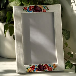 Może przedstawiać: Biała prostokątna ramka na zdjęcia z kolorowymi kwiatowymi akcentami. Ramka ma malowaną obwódkę z czerwonych, niebieskich, żółtych i fioletowych kwiatów. Ramka opiera się o białą powierzchnię, z zielonymi liśćmi w tle.