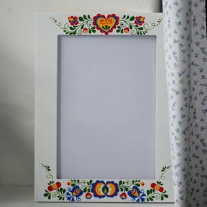 Könnte beinhalten: Weißer rechteckiger Bilderrahmen mit farbenfrohem Blumenmuster im Folk-Art-Stil. Der Rahmen hat eine zentrale Öffnung für ein Foto, verziert mit bemalten Blumen, Herzen und Vögeln in Rot, Gelb, Blau und Grün.
