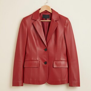 May include: A red leather blazer with a notched collar, two front pockets, and two black buttons. The blazer is hanging on a wooden hanger against a neutral background. A classic, tailored jacket.