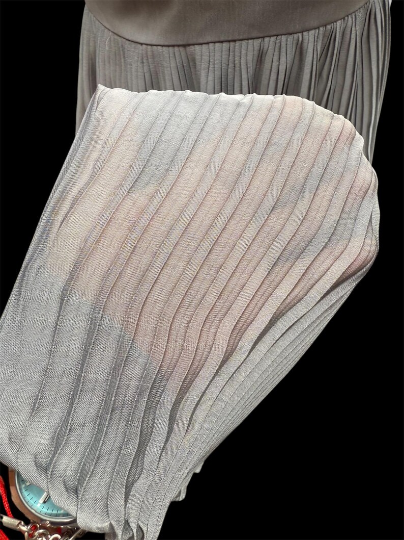 May include: A close-up of a gray pleated skirt made of a sheer fabric. The pleats are evenly spaced and create a delicate, flowing look.