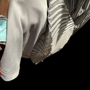 May include: A gray pleated fabric with a smooth texture. The fabric is folded and draped over a hand wearing a silver watch.