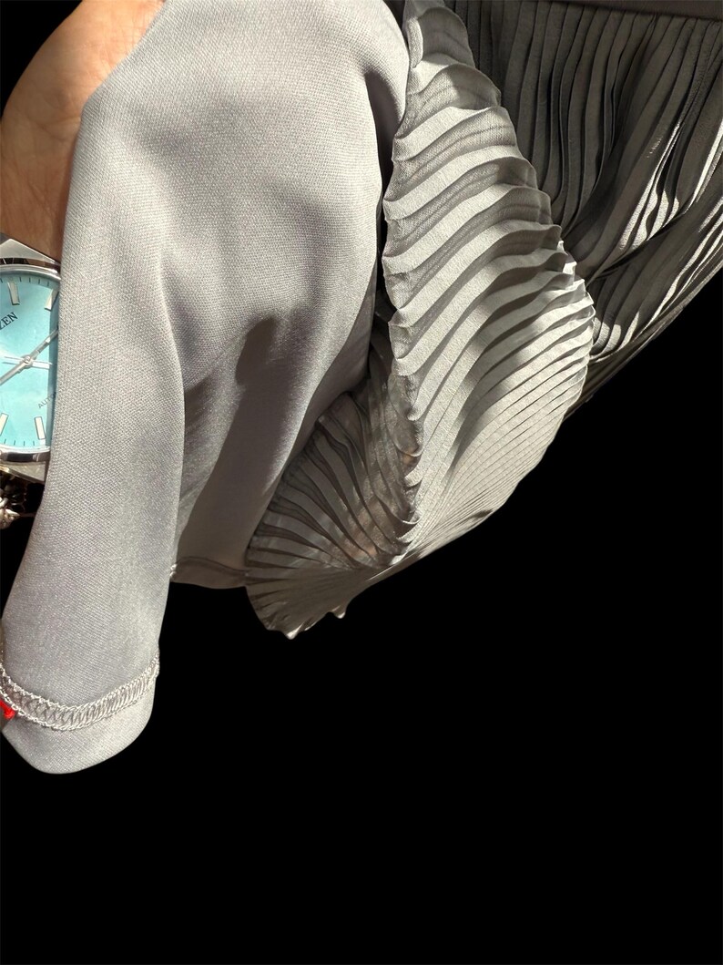 May include: A gray pleated fabric with a smooth texture. The fabric is folded and draped over a hand wearing a silver watch.