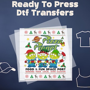 May include: A DTF transfer featuring the text "Ready To Press Dtf Transfers". The design includes three green aliens from Pizza Planet, a planet, and the text "Pizza Planet". The design is in a Christmas sweater style.